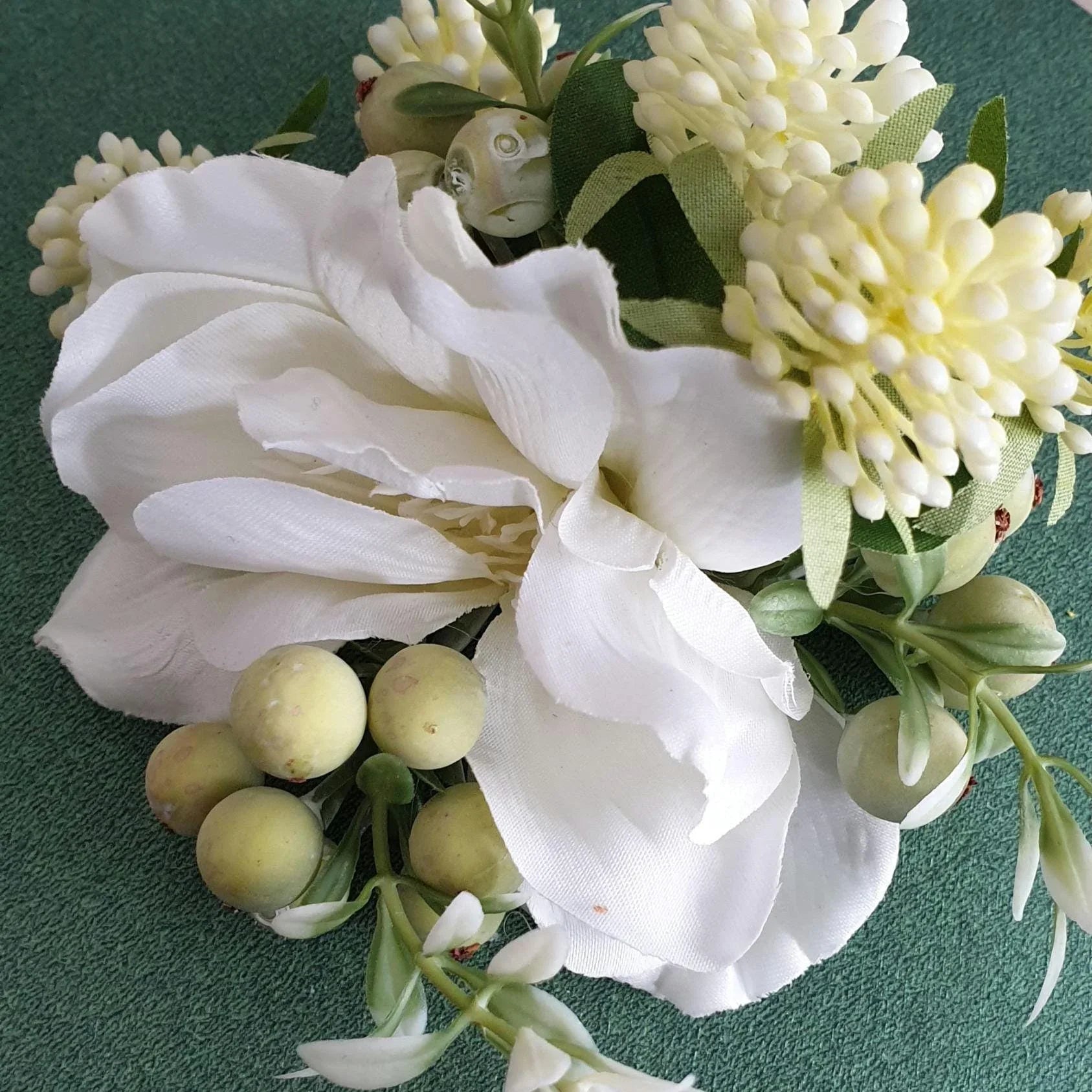 Artificial flower arrangement with white flowers, green leaves, and small yellow berries on a green background.