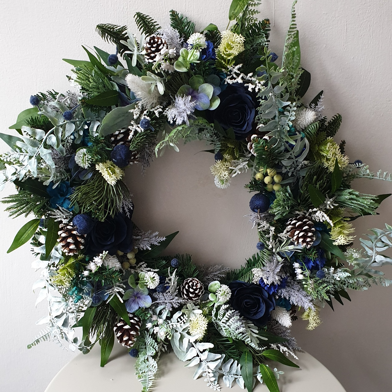 artificial wedding flowers - Decorative wreath with greenery, flowers, and pinecones on a white surface.