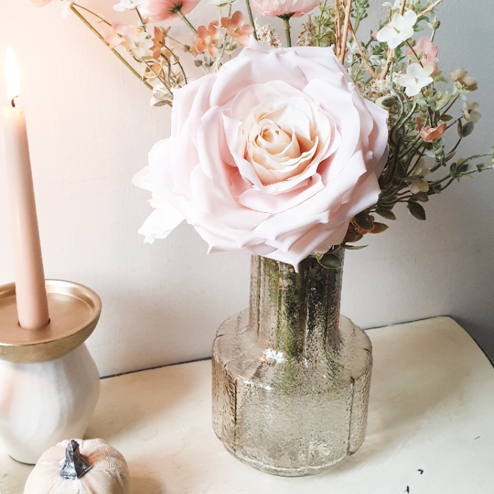 artificial wedding flowers - Pink rose in a glass vase on a white surface with a candle and flowers in the background.