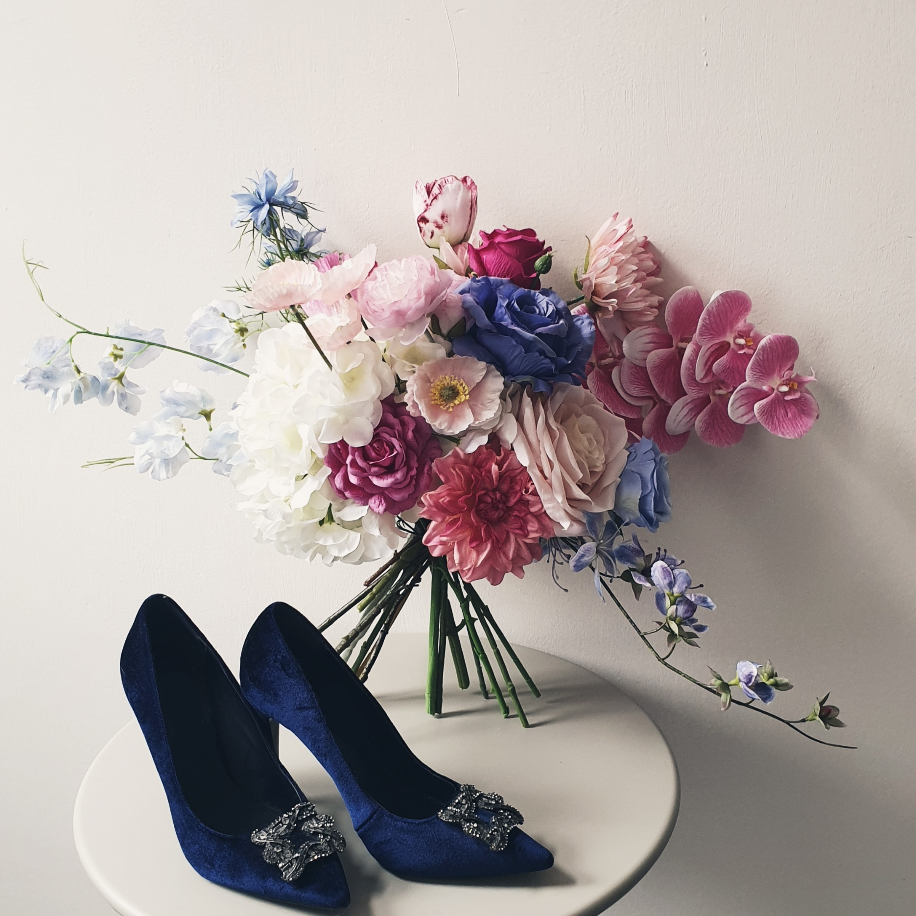 artificial wedding flowers - Pair of blue high-heeled shoes with decorative clips next to a bouquet of flowers on a white surface.
